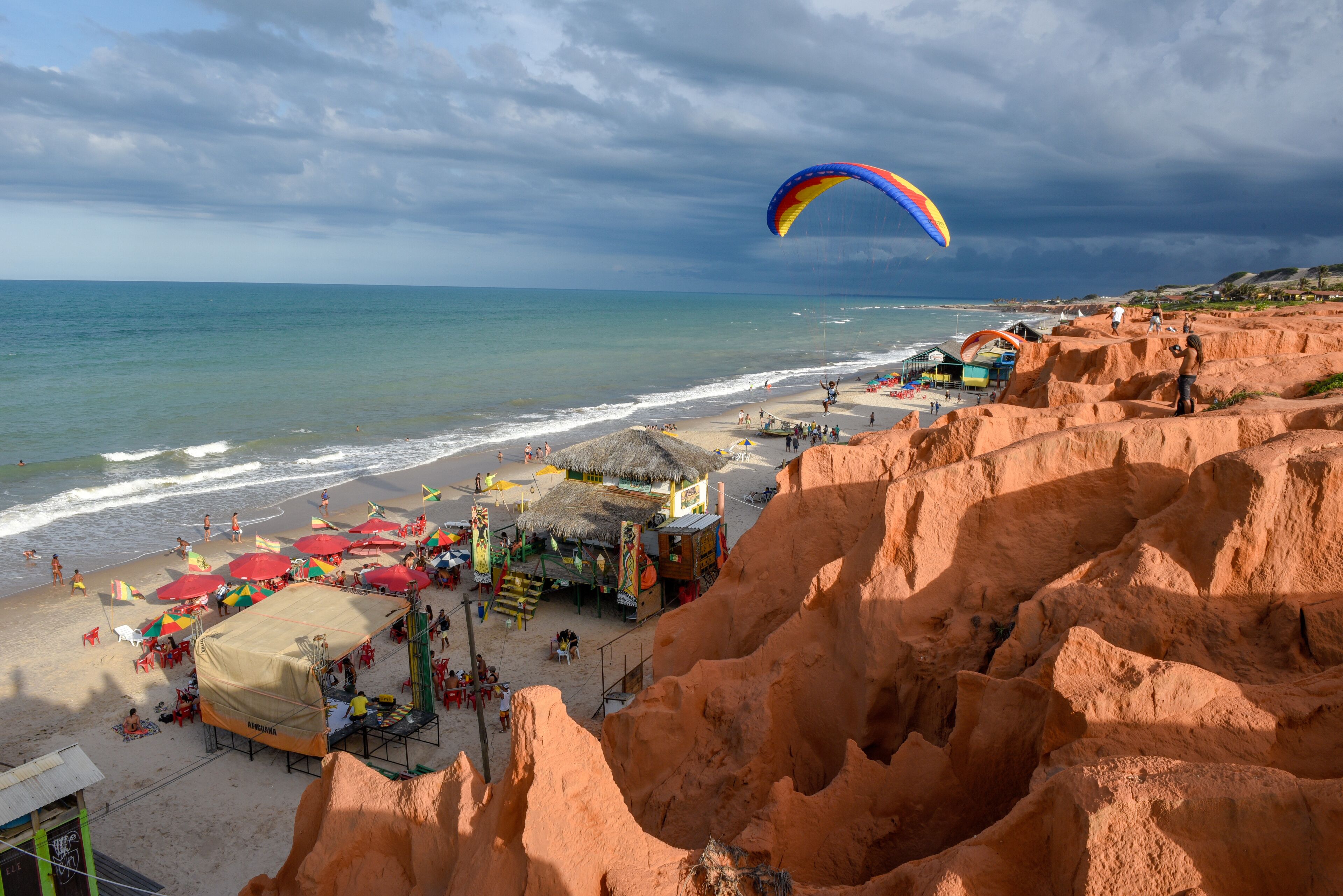 A beach with a parasail, a thatched-roof hut, and a rocky cliff.