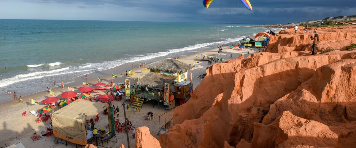 A beach with a parasail, a thatched-roof hut, and a rocky cliff.