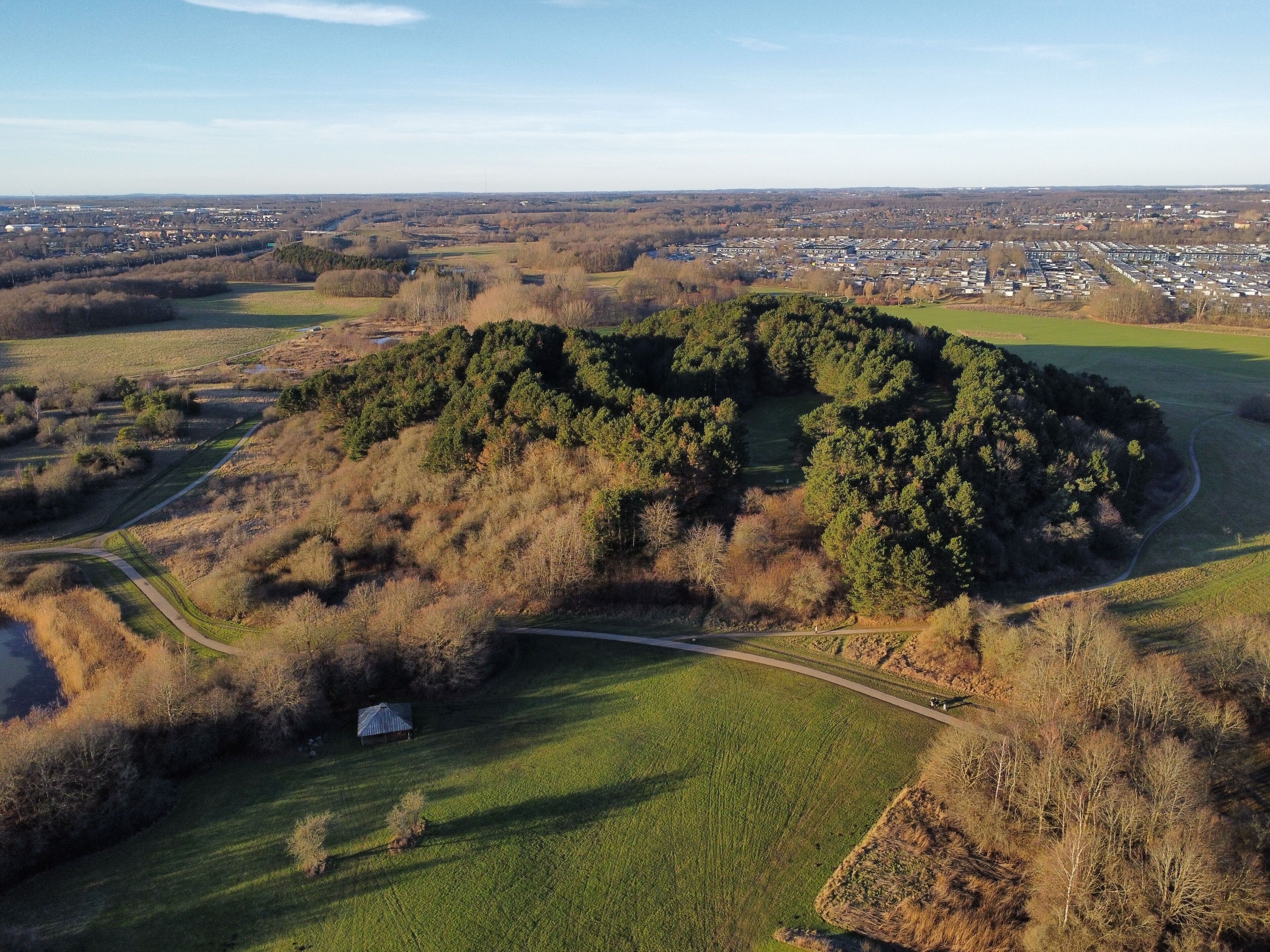 An aerial view of a forested hill with a small building, surrounded by open fields and a distant town.