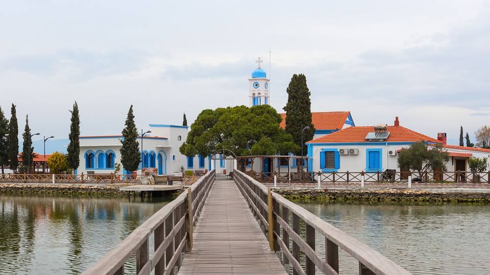 A wooden pier leading to a church with blue and white buildings, a bell tower, and a calm lake.
