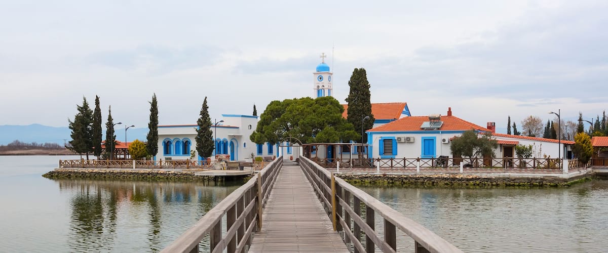 A wooden pier leading to a church with blue and white buildings, a bell tower, and a calm lake.