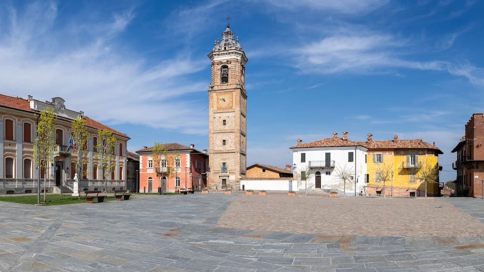 A historic square with a prominent clock tower, surrounded by colorful buildings and a paved plaza.
