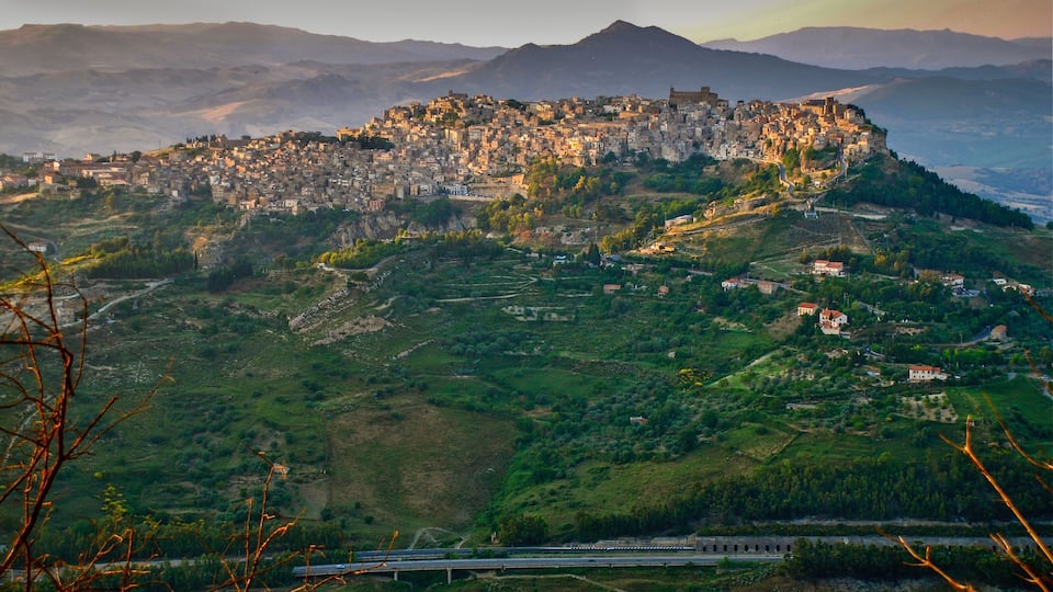 A mountainous town with a castle on a hill, surrounded by greenery and a railway track.