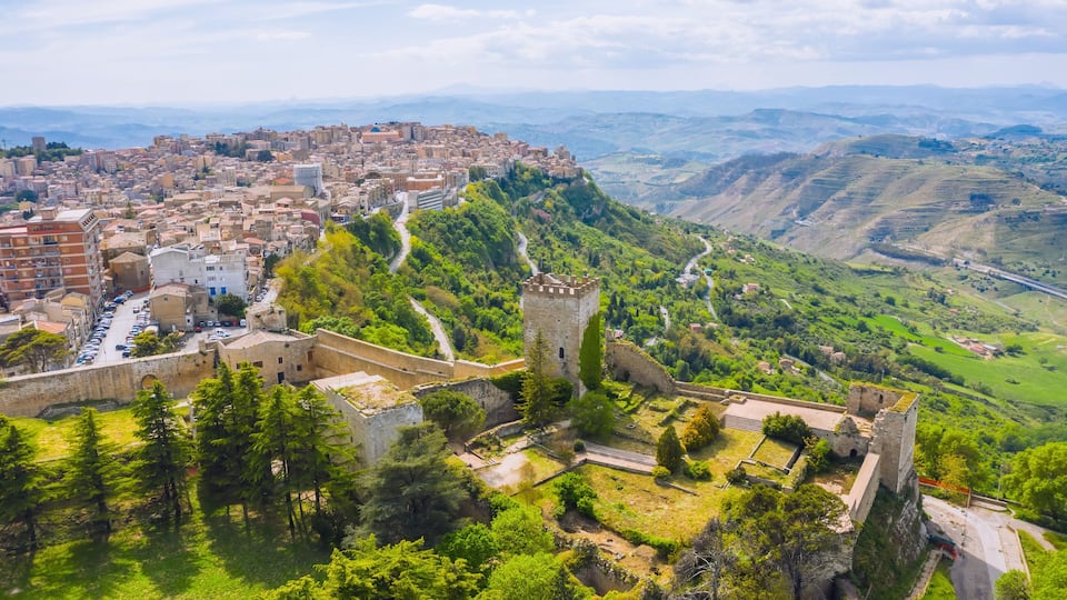 A cityscape with a historic fortress on a hill, surrounded by greenery and a valley.
