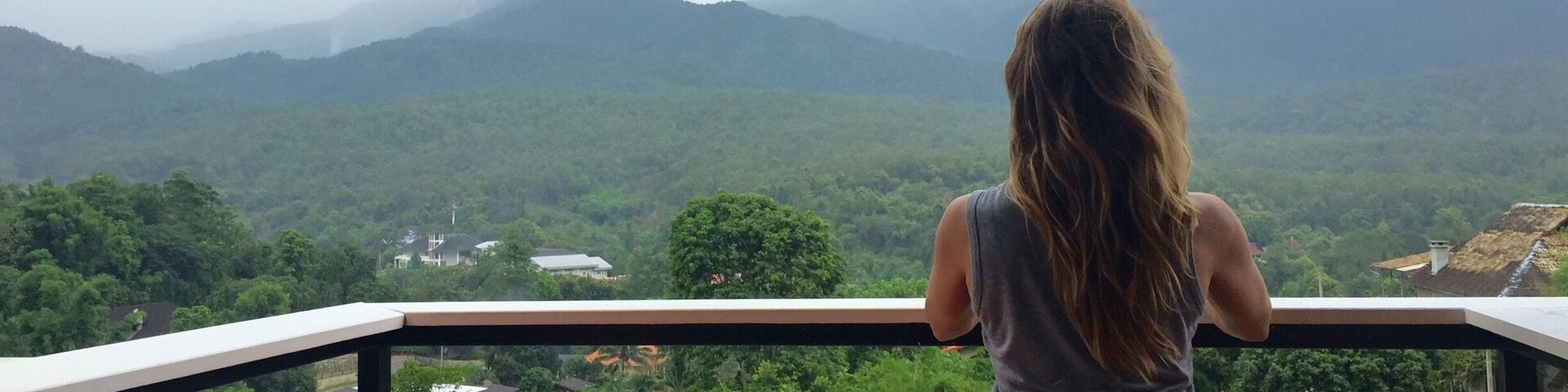 Una persona sentada en un balcón con vista a un paisaje montañoso, con vegetación y casas.