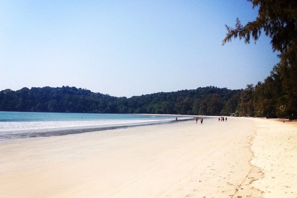A sandy beach with a clear blue sky and a forested hill in the background.
