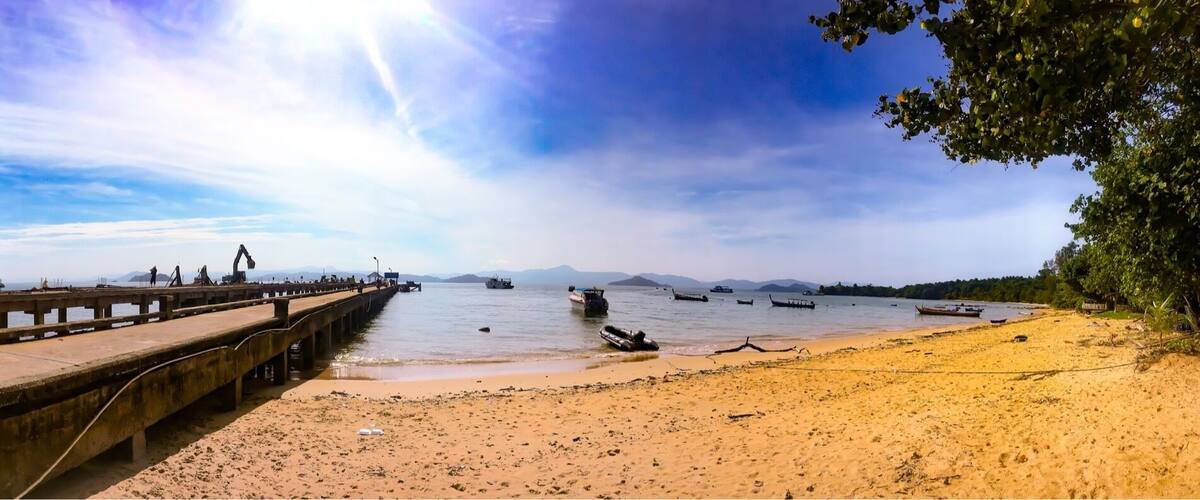 A pier extending into calm waters with boats, a sandy beach, and distant mountains.