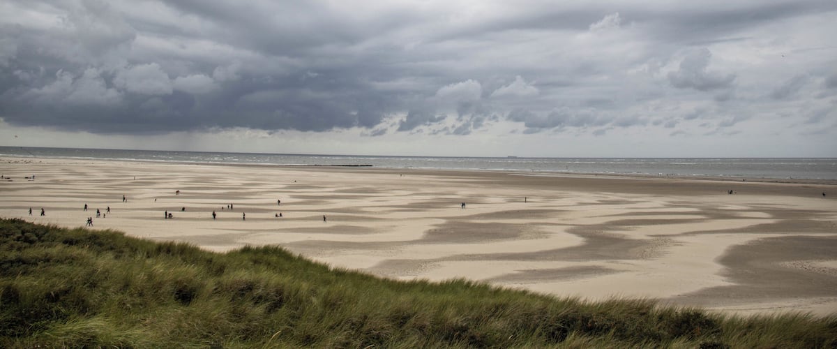 A beach with people walking, overcast sky, and grassy dunes.