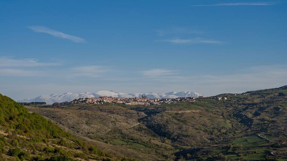 A mountainous landscape with a town nestled among the hills, snow-capped peaks in the distance, and a clear blue sky.