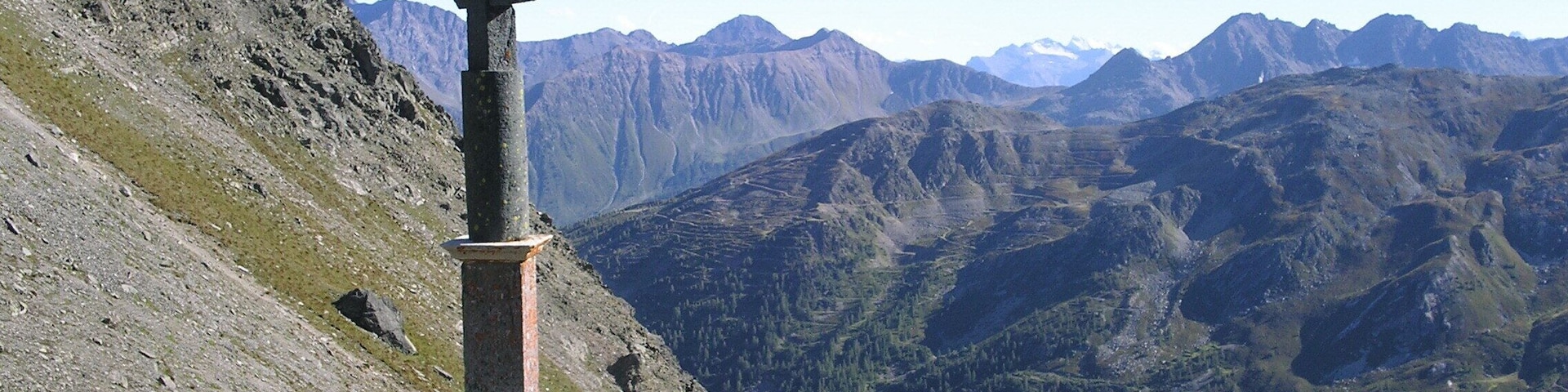 Una cruz de piedra en la cresta de una montaña, con un valle y montañas lejanas al fondo.