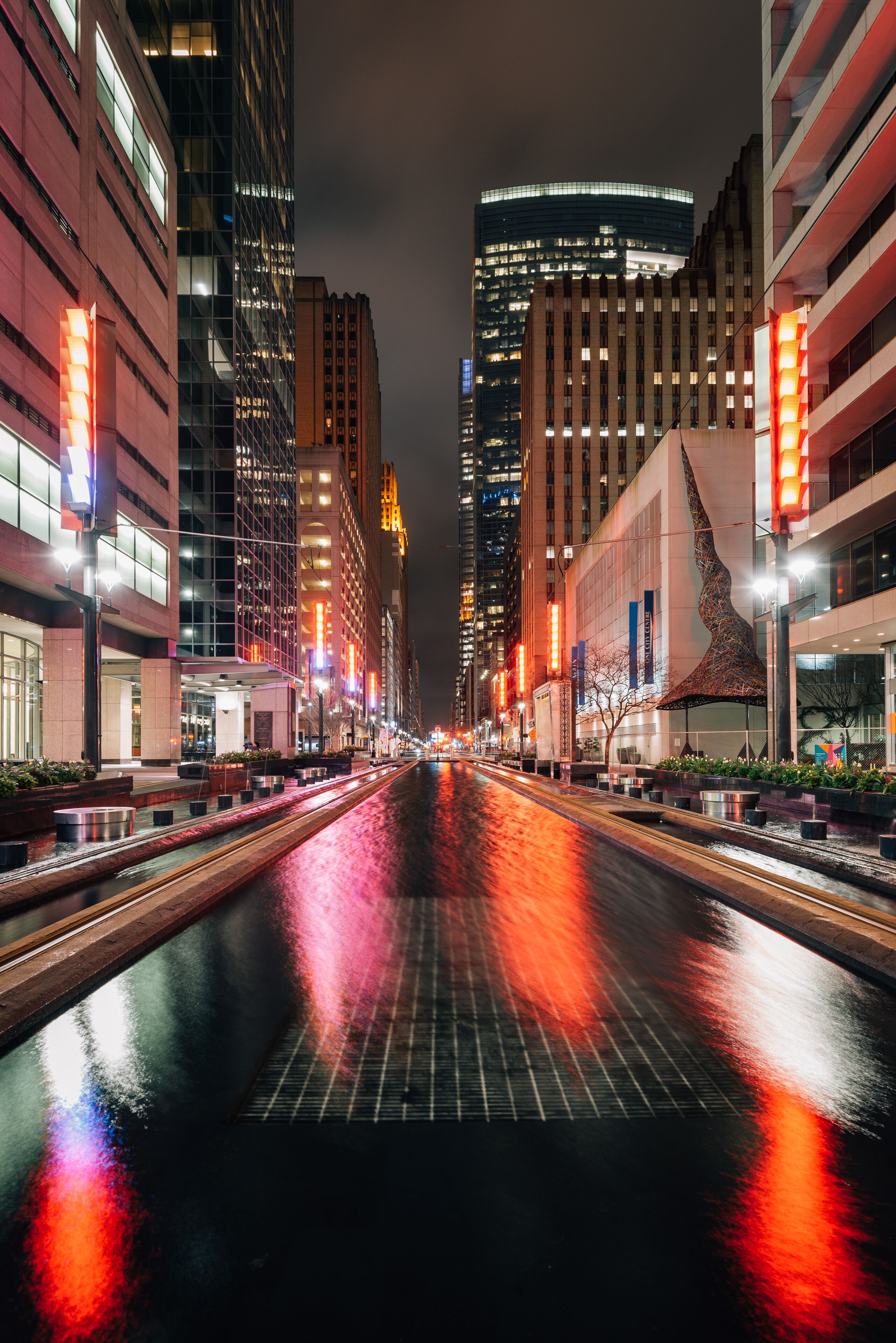 A nighttime city street with illuminated buildings and a wet road reflecting lights.