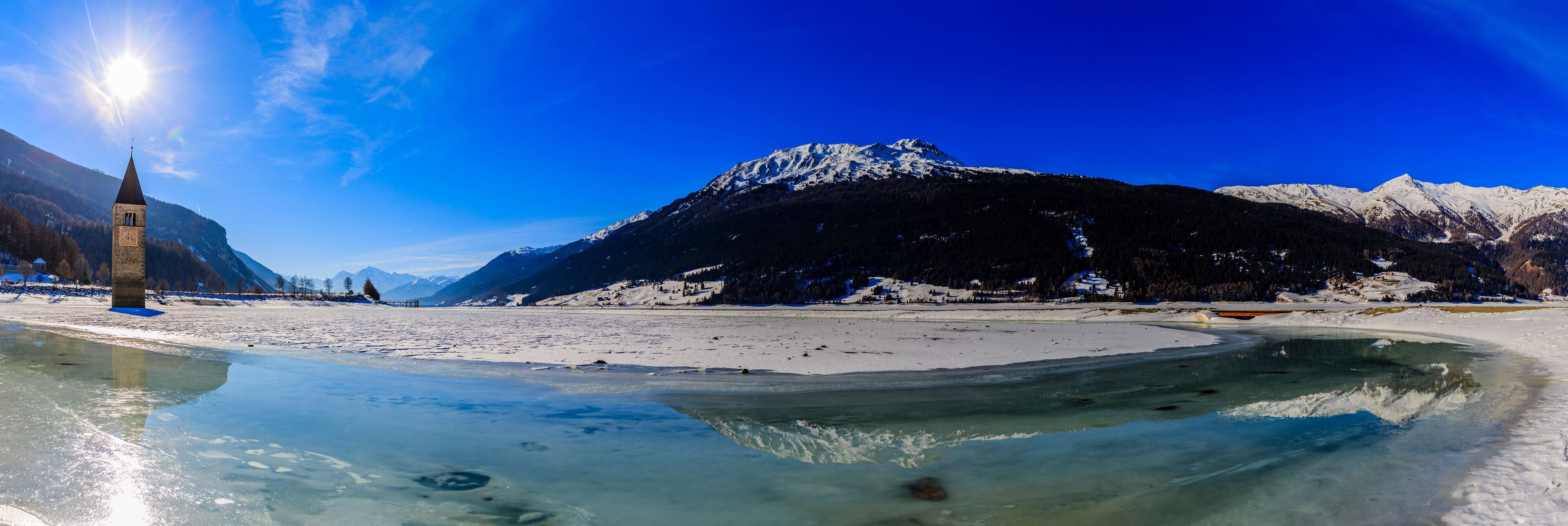 A frozen lake with a church spire, snow-covered mountains, and clear blue sky.