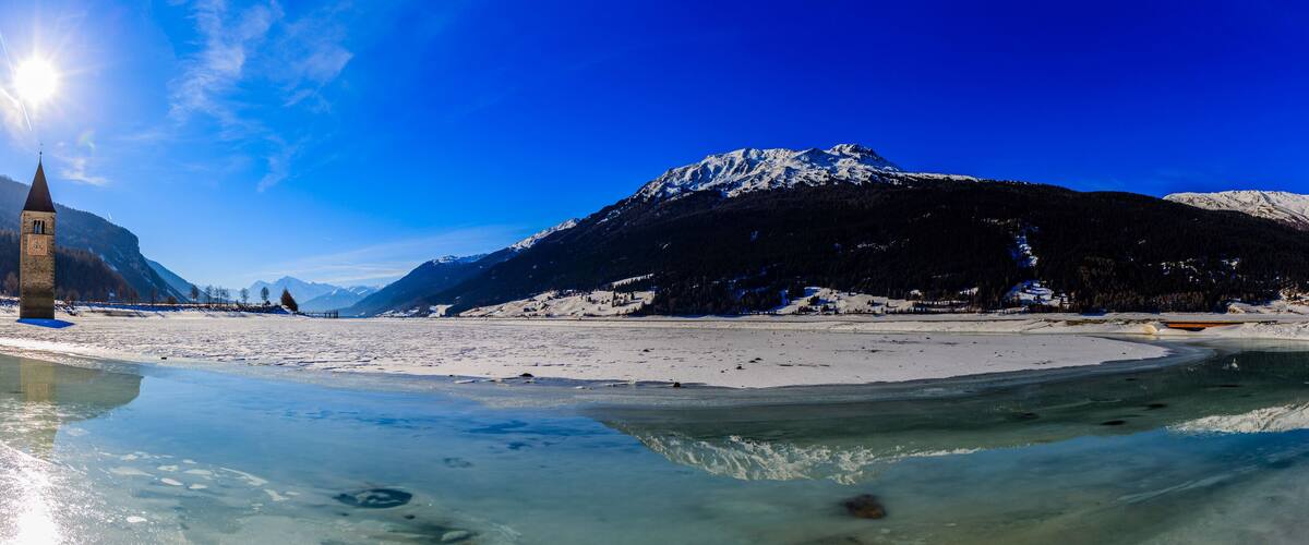 A frozen lake with a church spire, snow-covered mountains, and clear blue sky.