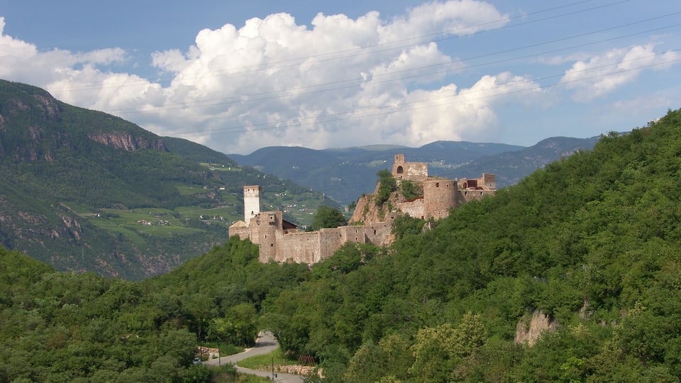 A historic castle on a hill with a winding road leading to it, surrounded by lush greenery and mountains.