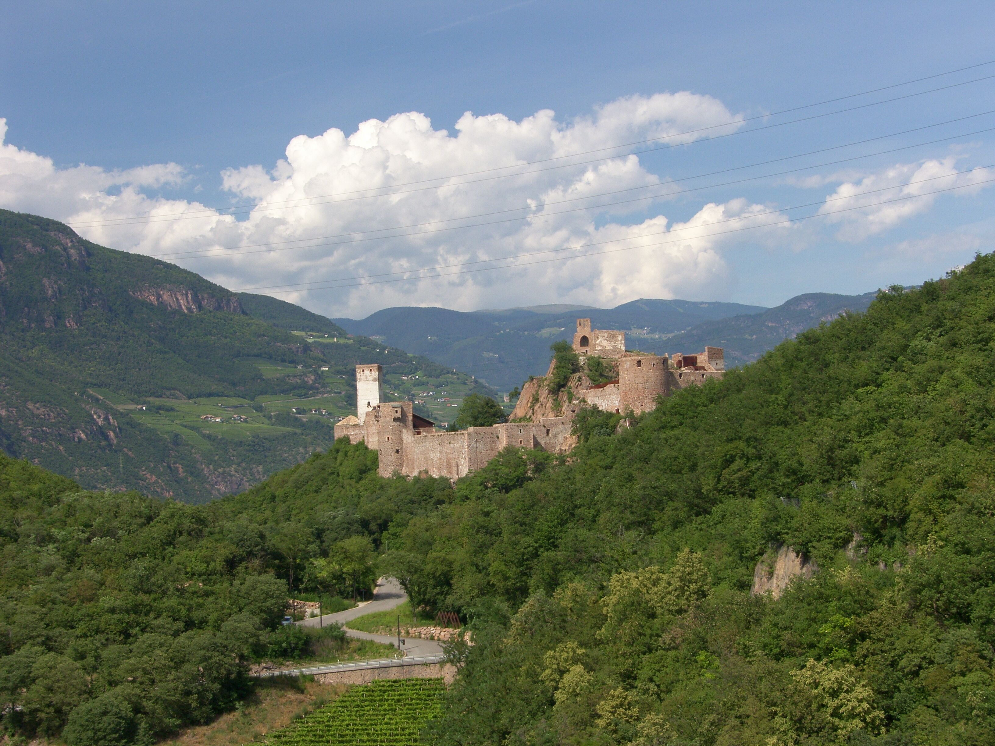 A historic castle on a hill with a winding road leading to it, surrounded by lush greenery and mountains.