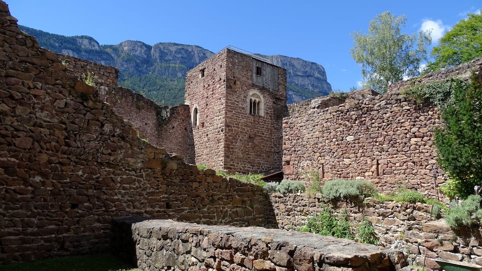 An ancient stone structure with a tower, surrounded by mountains and greenery.