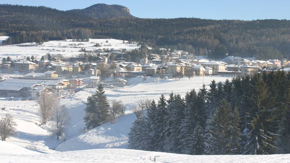 A snow-covered village with buildings and a church, surrounded by forested hills.