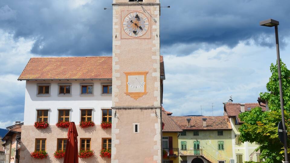 A historic clock tower with a pointed roof, surrounded by traditional buildings with red flower boxes and a cobblestone street.