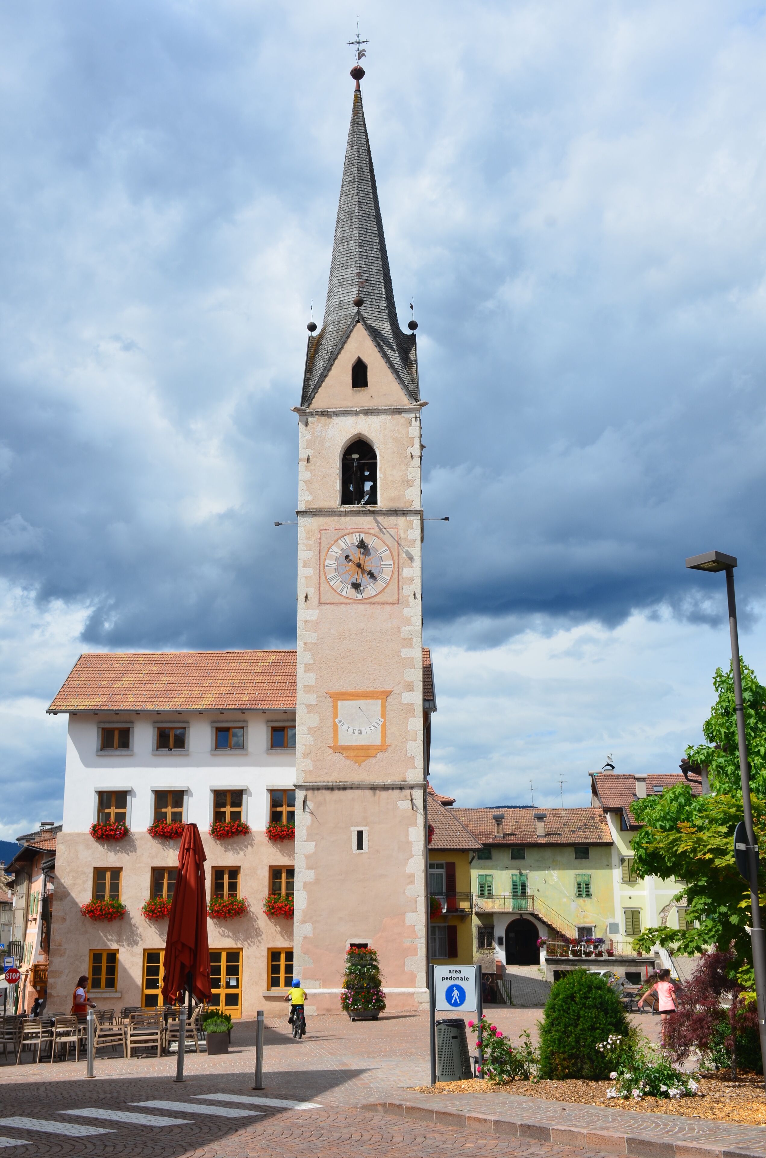 A historic clock tower with a pointed roof, surrounded by traditional buildings with red flower boxes and a cobblestone street.