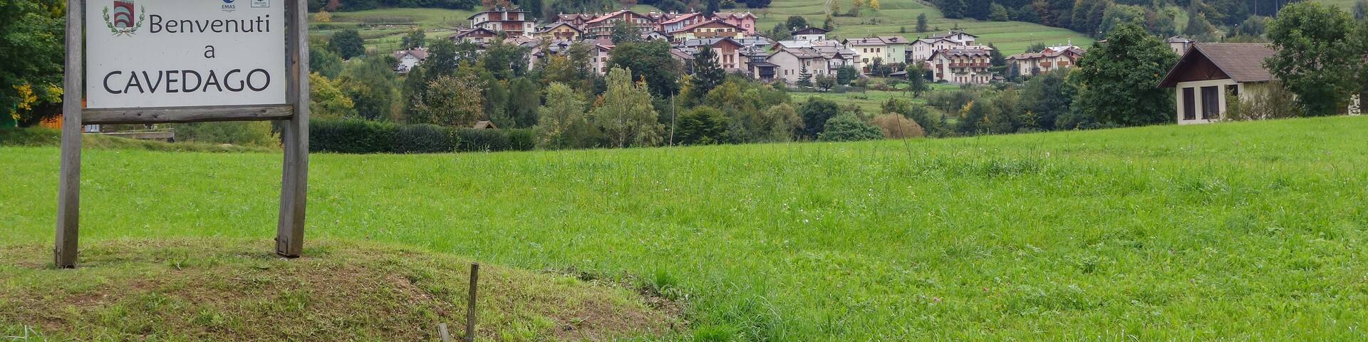 A sign reading "Benvenuti a CAVEDAGO" with a village in the background surrounded by mountains.
