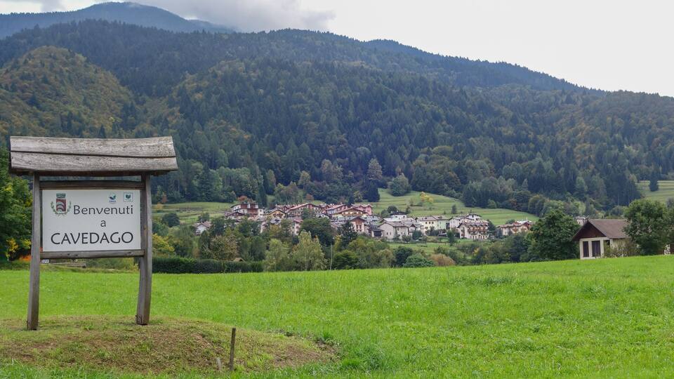 A sign reading "Benvenuti a CAVEDAGO" with a village in the background surrounded by mountains.