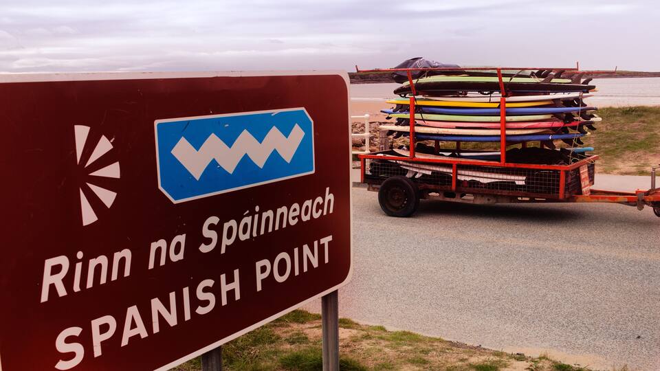 A sign reading "Rinn na Spanish Point" with a cart of kayaks in the background.