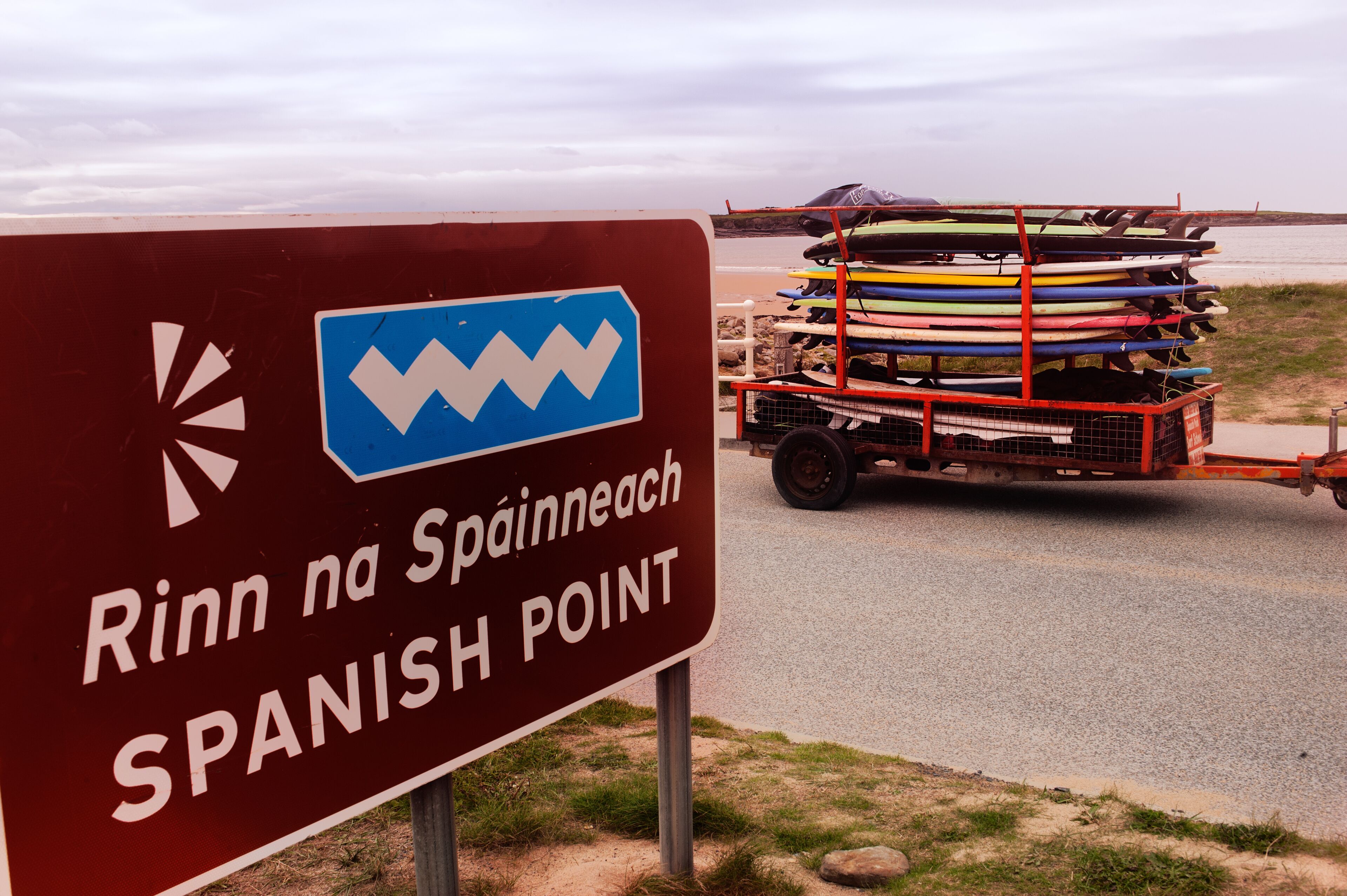 A sign reading "Rinn na Spanish Point" with a cart of kayaks in the background.