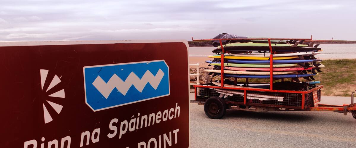 A sign reading "Rinn na Spanish Point" with a cart of kayaks in the background.