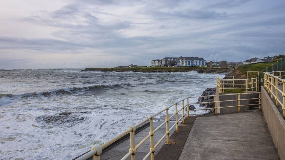 A coastal walkway with a railing, waves crashing against the shore, and a row of houses in the background.