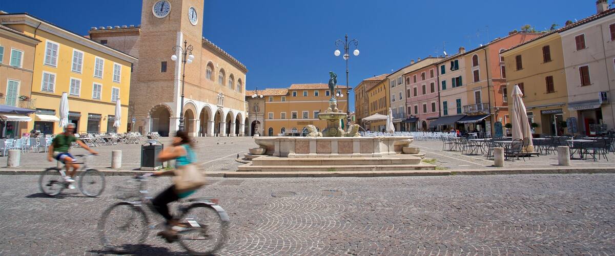A cobblestone square with a historic tower, a fountain, and outdoor seating areas.