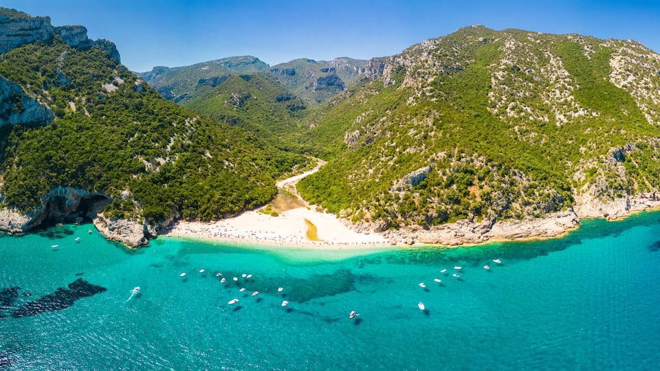 A mountainous island with a beach, clear turquoise waters, and boats.