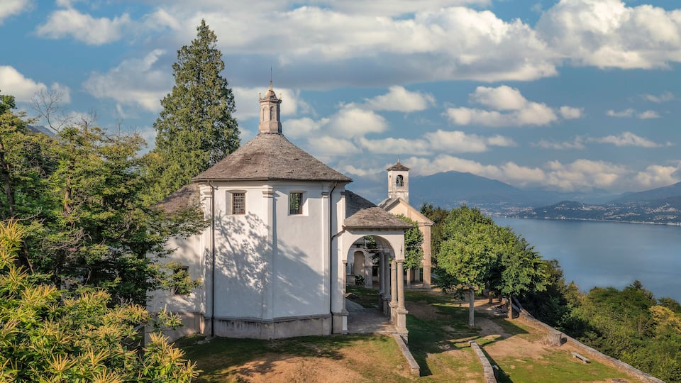 A white church with a bell tower and a small tower, surrounded by trees and a lake.