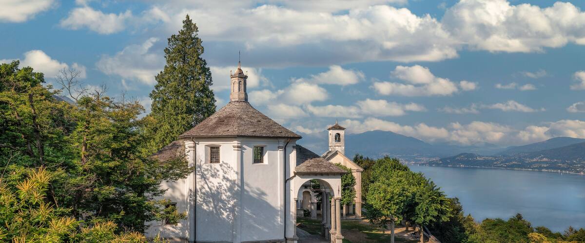 A white church with a bell tower and a small tower, surrounded by trees and a lake.
