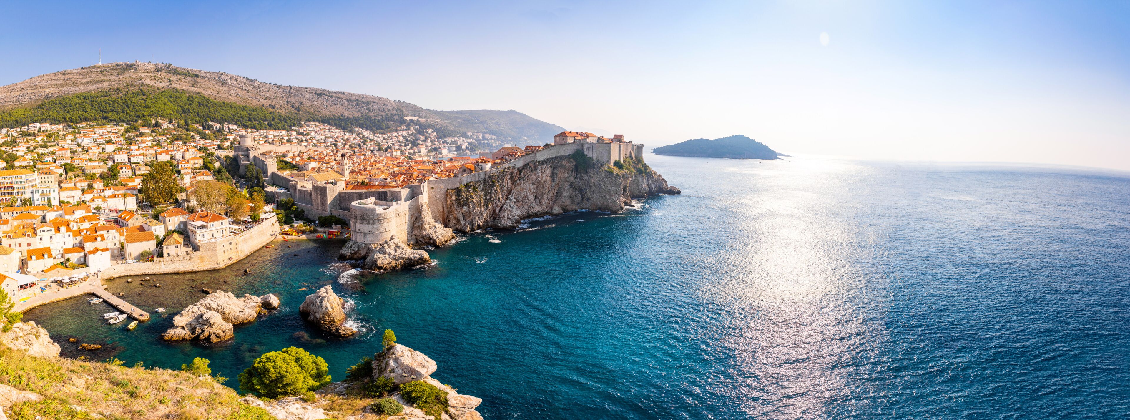 A coastal town with a historic fortification, a clear blue sea, and a hilly backdrop.