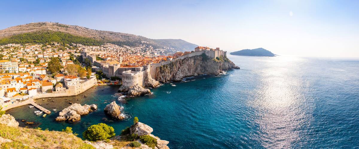 A coastal town with a historic fortification, a clear blue sea, and a hilly backdrop.