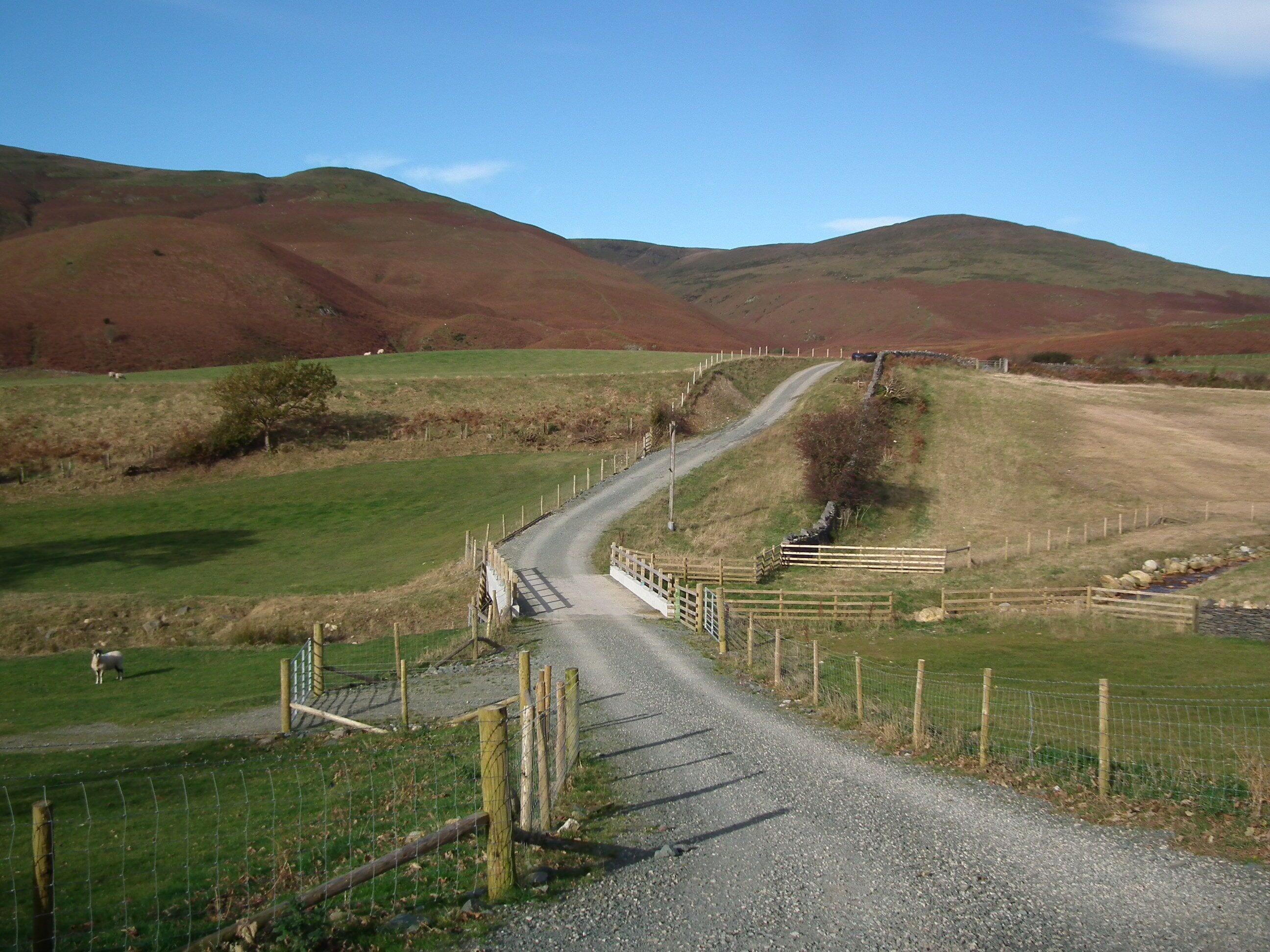 Uma estrada de cascalho serpenteia por uma paisagem acidentada, com campos verdes e um muro de pedra à direita.