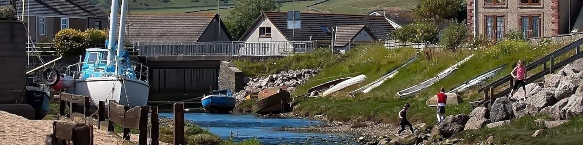 A coastal village with boats, a river, and houses with a hill in the background.