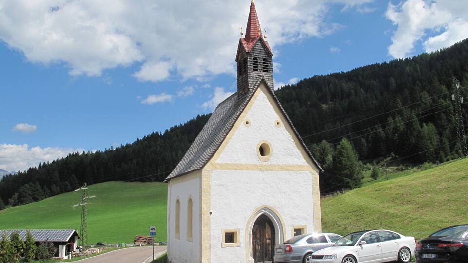 A small church with a red roof and white walls, parked cars in front, and a green hillside in the background.