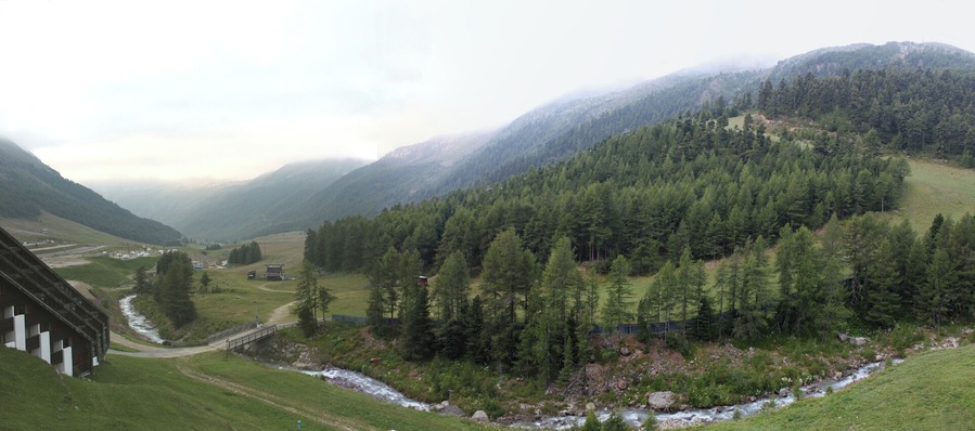 Paisaje montañoso con río, bosque denso y un edificio a la izquierda.