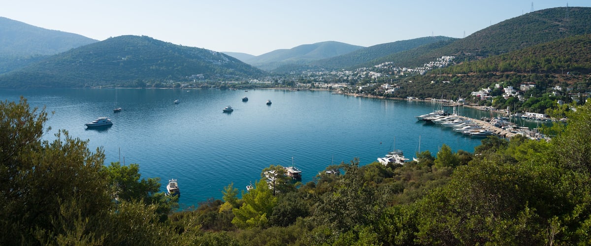 A coastal town with boats docked along the shoreline, surrounded by hills and clear blue water.