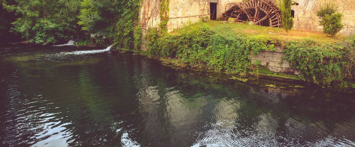 A historic building with a water wheel, surrounded by lush greenery and a calm river.
