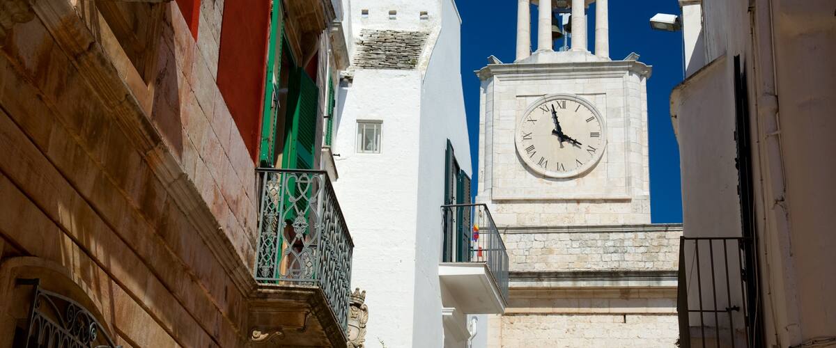 A clock tower with a dome and a bell, surrounded by buildings with balconies and a clear blue sky.