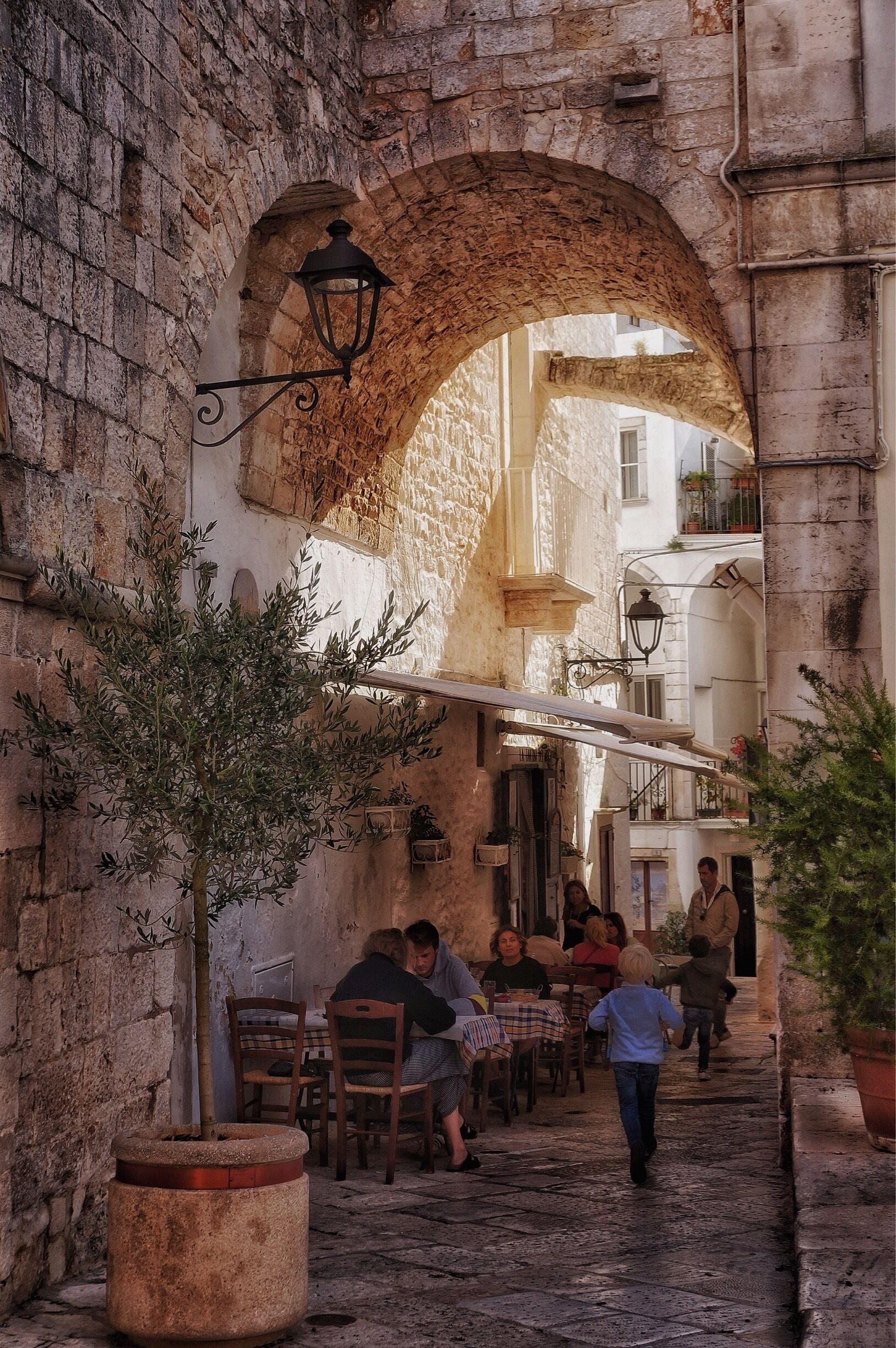 A narrow cobblestone street with stone buildings, a large archway, and outdoor seating at a café.