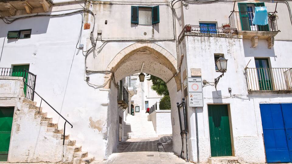 A narrow street with a stone archway, white buildings with green shutters, and a blue door.