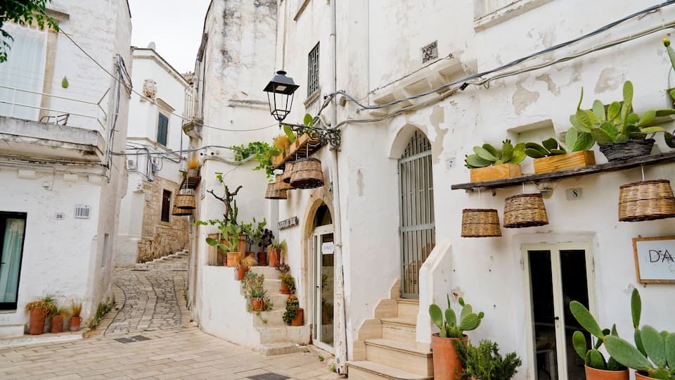 A narrow cobblestone street flanked by white buildings with potted plants and wicker baskets.