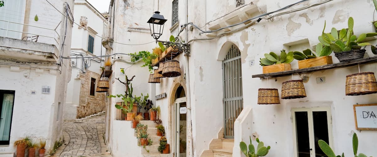 A narrow cobblestone street flanked by white buildings with potted plants and wicker baskets.