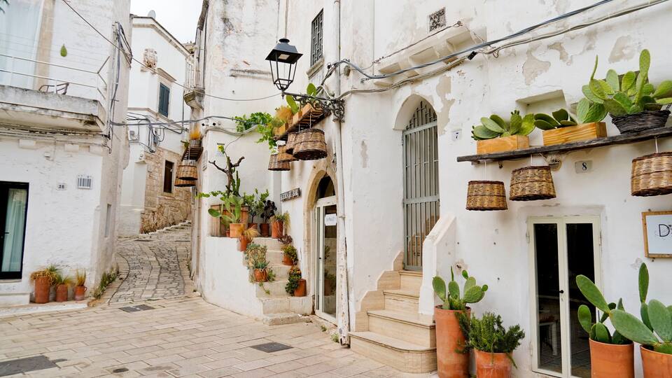 A narrow street with white buildings, potted plants, and a staircase leading to a bar.