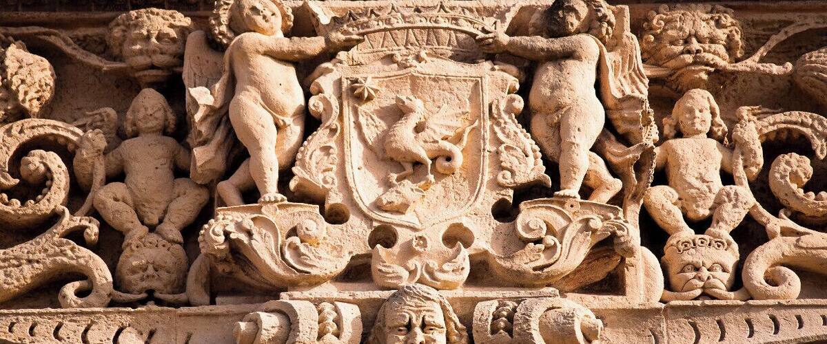 A detailed stone carving with a central shield emblem, flanked by figures and intricate designs.