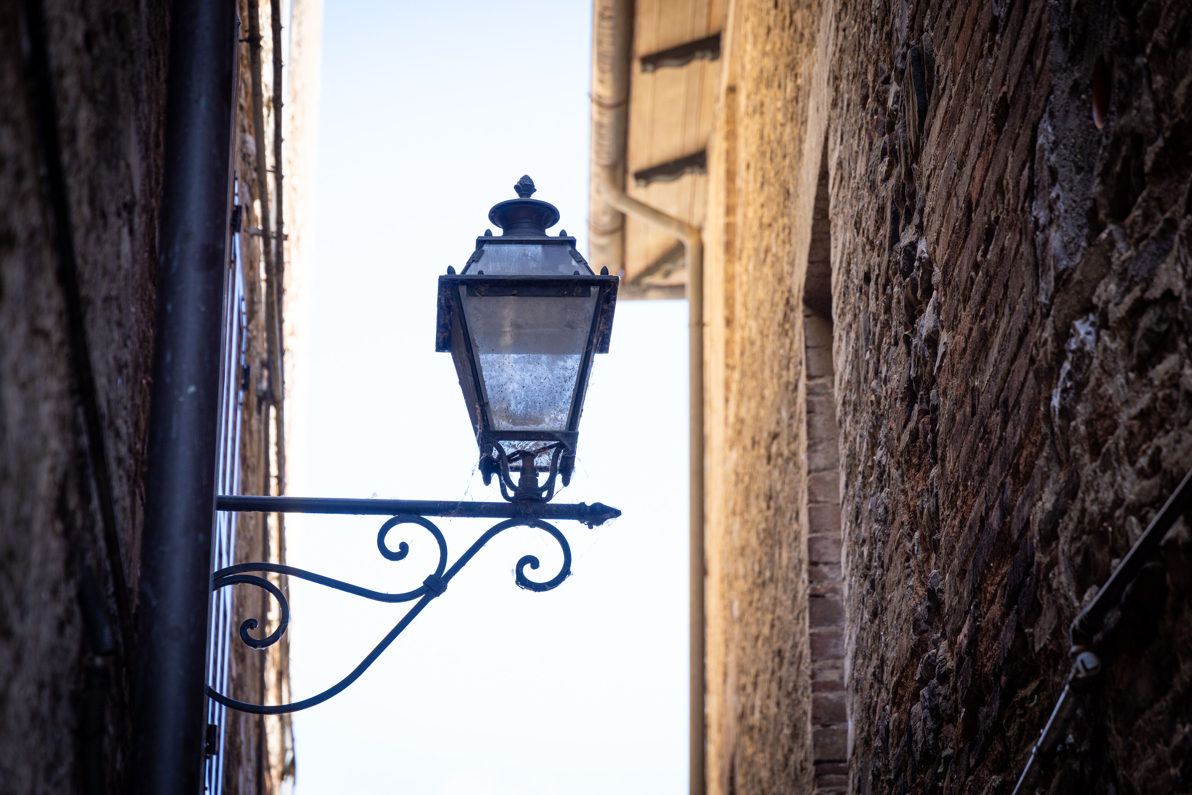 A street lamp attached to a metal pole with a decorative scroll design, set against a backdrop of aged brick buildings.