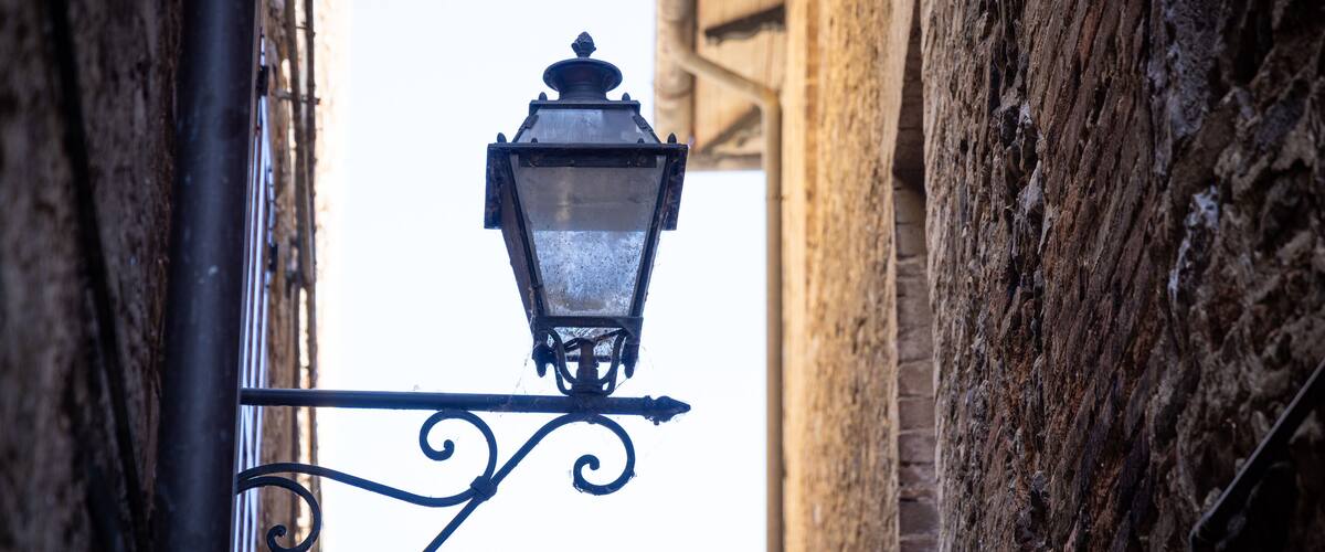 A street lamp attached to a metal pole with a decorative scroll design, set against a backdrop of aged brick buildings.